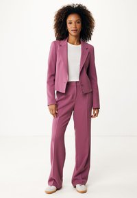 Blazer ajusté rose avec un pantalon large assorti. Le blazer a un design à un bouton et une texture subtile. Porté sur une chemise blanche.