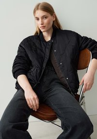 Blouson aviateur noir matelassé avec fermeture éclair, poignets côtelés et doublure lisse, associé à un jean droit sombre. Modèle assis sur une chaise.