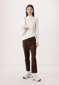 Hvit turtleneck-genser, brune bukser og sølvfargede sneakers. Strukturert hvit veske med et mønstrete bånd. Modellen står mot en nøytral bakgrunn.