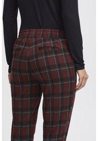 Pantaloni a scacchi marroni con accenti neri e verdi, dotati di una vita elastica e tasca posteriore, mostrati da dietro su uno sfondo chiaro.