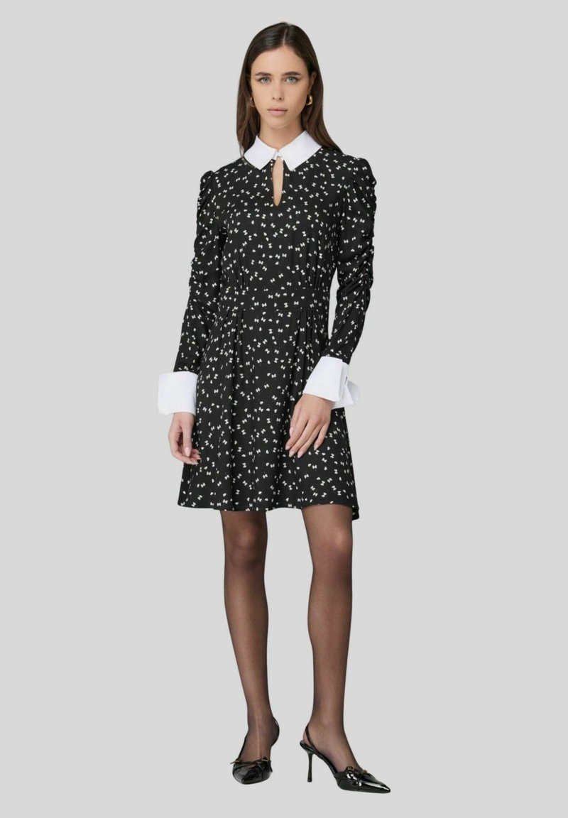 Abito nero con motivo floreale bianco, maniche lunghe, scollatura a chiave e colletto e polsini bianchi a contrasto. Indossato con tacchi neri.