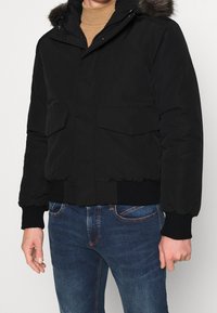 Man wearing a black puffer jacket with fur-trimmed hood, ribbed cuffs and hem, paired with blue jeans and a tan turtleneck.