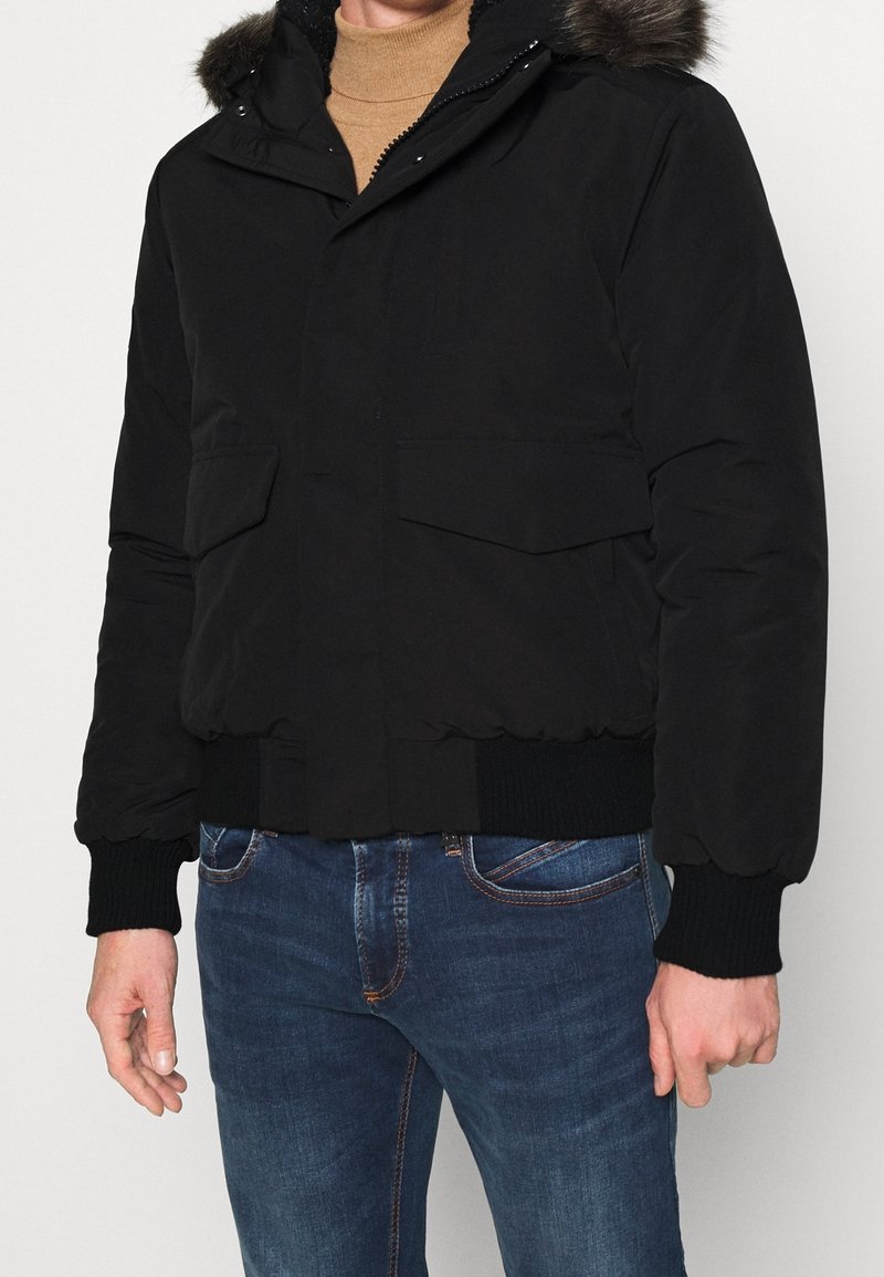 Man wearing a black puffer jacket with fur-trimmed hood, ribbed cuffs and hem, paired with blue jeans and a tan turtleneck.