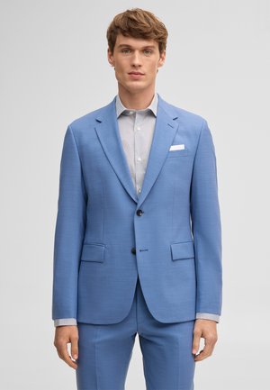 Mann in hellblauem Anzug und gemustertem weißen Hemd, der vor einem schlichten grauen Hintergrund steht und nach vorne schaut.