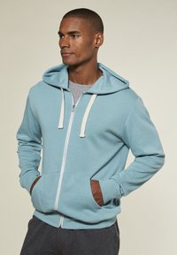 Hellblauer Reißverschluss-Hoodie mit einer verstellbaren Kapuze, kontrastierenden beigen Kordeln und braunen Akzenten an den Fronttaschen.