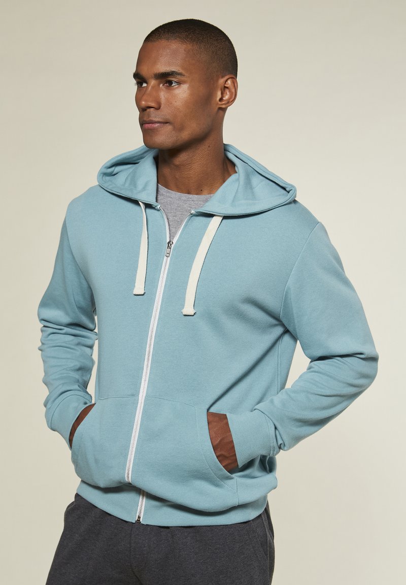 Hellblauer Reißverschluss-Hoodie mit einer verstellbaren Kapuze, kontrastierenden beigen Kordeln und braunen Akzenten an den Fronttaschen.