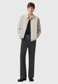 Lichtbeige bomberjack met een gladde textuur, gecombineerd met losse zwarte denim jeans. Model staat met de handen in de zakken en draagt een zwart shirt.