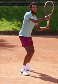 Tennisser in een lichtblauw shirt en kastanjebruine shorts, met een racket in de hand, gekleed in witte schoenen met zwarte accenten, op een gravelbaan.