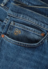 Jeans de mezclilla en azul oscuro con costuras naranjas en contraste. Cuenta con un bolsillo frontal con un logo de oso bordado en beige.