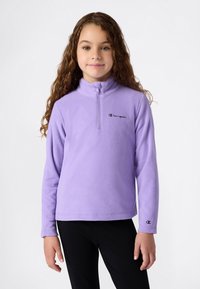 Fille avec de longs cheveux bouclés portant un pull en polaire violet Champion avec demi-fermeture éclair et un pantalon noir, se tenant devant un fond uni.