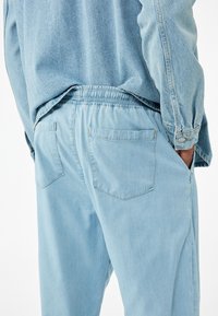 Lichtblauwe denim broek heeft een elastische tailleband, twee achterzakken en een relaxte pasvorm, gecombineerd met een lichtblauwe denim shirt.