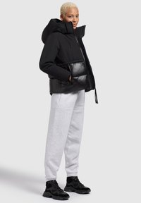 Zwarte capuchon pufferjack met een glanzend zwart onderste gedeelte, gecombineerd met grijze sweatpants en zwarte chunky sneakers.