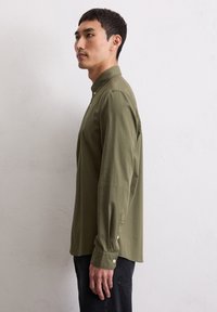 Olijfgroene, lange mouwen overhemd met een button-down kraag. Voorzien van manchetknoopjes en een rechte zoom. Gedragen met zwarte broek.