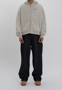 Krämfärgad zip-up-hoodie med framficka, kombinerad med löst sittande mörk denimjeans och bruna stövlar. Enkel design utan synliga mönster.