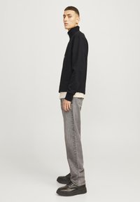 Sudadera negra con cremallera y cuello alto, usada sobre una camisa crema, combinada con jeans grises y zapatos negros, mostrada de perfil.