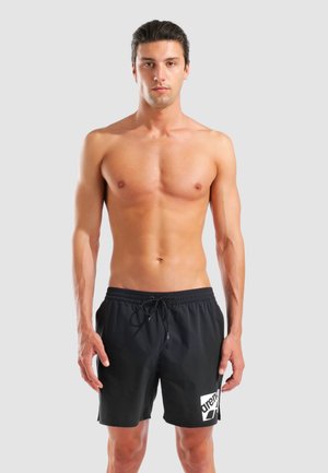 Uomo senza maglietta con capelli corti e scuri che indossa pantaloncini da bagno neri con il logo bianco "arena", in piedi contro uno sfondo chiaro uniforme.