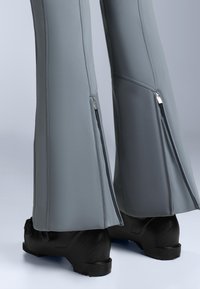 Grå vidbenta byxor gjorda av flexibelt material med metallblixtlås vid vaden. Svarta stövlar delvis synliga längst ner.