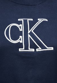 Ναυτικό μπλε πλεκτό ύφασμα με μεγάλα λευκά κεντημένα γράμματα "cK" στο κέντρο της περιοχής του στήθους.