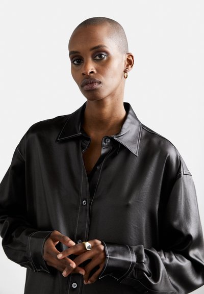 Black leather shirt with a classic collar, visible stitching accents, and buttons. Model wearing a geometric ring. Smooth texture.