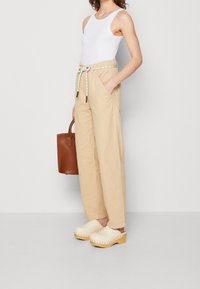 Bežové široké kalhoty s elastickým pasem a bočními kapsami, v kombinaci s bežovými dřevěnými klínovými botami a hnědou plátěnou taškou. Bílé tílko.