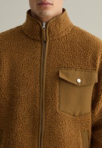 Bruine fleece jas met een hoge kraag, rits sluiting en een contraststof zak met een drukknoopdetail. Zachte, gestructureerde oppervlakte.