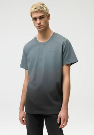 Młody mężczyzna o krótkich blond włosach, ubrany w koszulkę z krótkim rękawem w gradientowym kolorze od szarego do czarnego oraz ciemne spodnie, stojący na jednolitym tle.