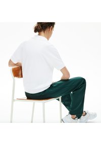 Vit kortärmad polo med ribbad krage, parat med mörkgröna avsmalnande byxor. Modellen sitter på en trästol och har på sig vita sneakers.