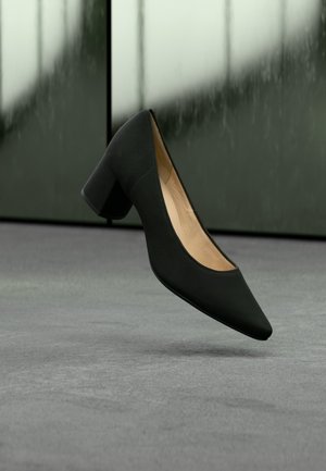 Sort damespumpe med spids tå og blokhæl svæver over en grå gulv med sløret grønt og sort baggrund.