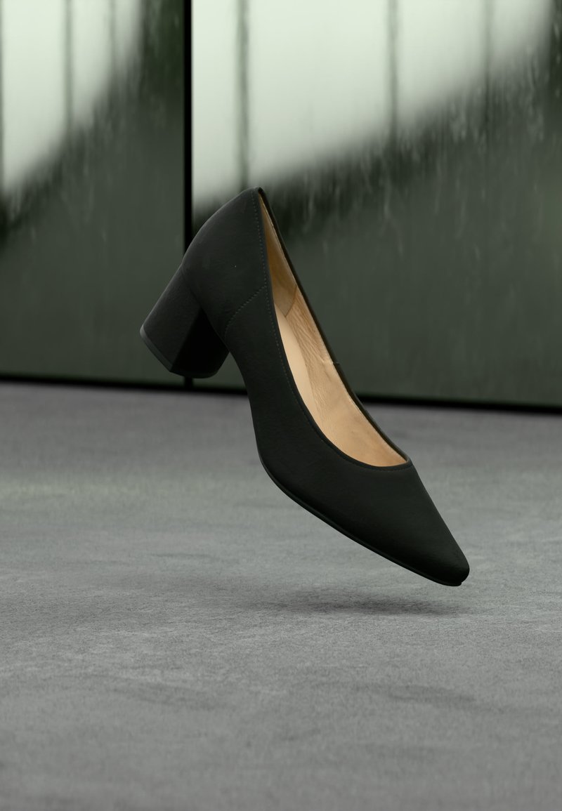Γυναικείο παπούτσι μαύρου χρώματος με μυτερή μύτη και τακούνι block, να αιωρείται πάνω από γκρι πάτωμα με θολό πράσινο και μαύρο φόντο.