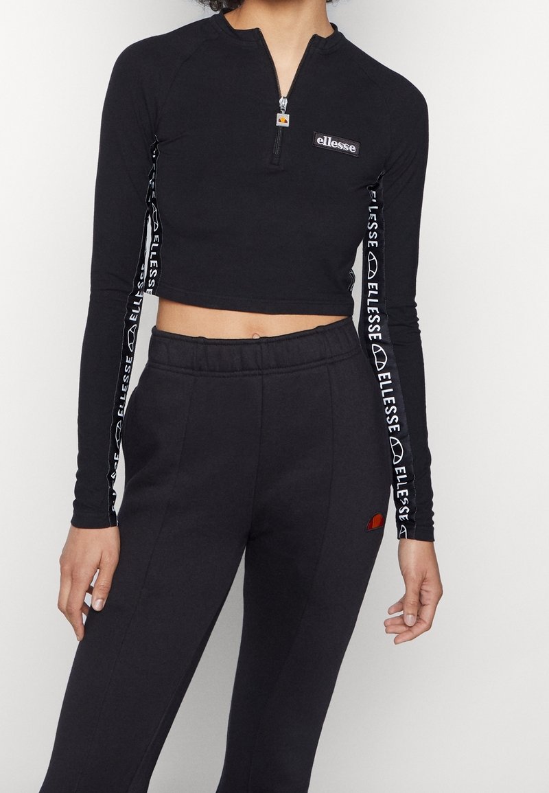 Black cropped long-sleeve top with a zip neck, featuring white logo tape on sleeves. Paired with black elastic waist sweatpants with logo.