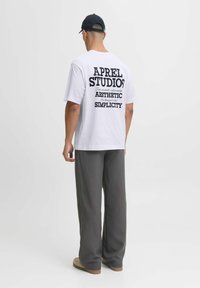 Valkoinen t-paita, jonka selässä on suurta mustaa tekstiä, jossa lukee "APREL STUDIOS," "AESTHETIC" ja "SIMPLICITY." Yhdistetty löysiin harmaisiin housuihin ja beigenvärisiin kenkiin.