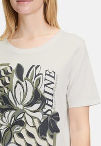 T-shirt beige clair à manches courtes avec un imprimé floral en vert, noir et blanc, portant le texte "SHINE" et des accents de motif en vagues. Matière en coton.