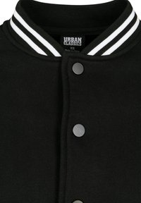 Veste de style varsity noire avec un col en rib à deux rayures blanches. Elle possède des boutons pression noirs et une texture douce. Étiquette de marque visible à l'intérieur.