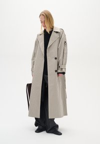Trench coat lungo, di colore chiaro, con un motivo a quadri, colli oversize, bottoni doppi e una vestibilità rilassata. Maglione e pantaloni neri.