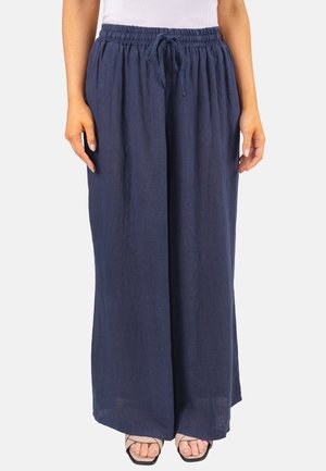 Femme portant un pantalon large ample bleu marine avec une taille élastique et un cordon de serrage, associé à un haut blanc et des sandales noires à lanières.