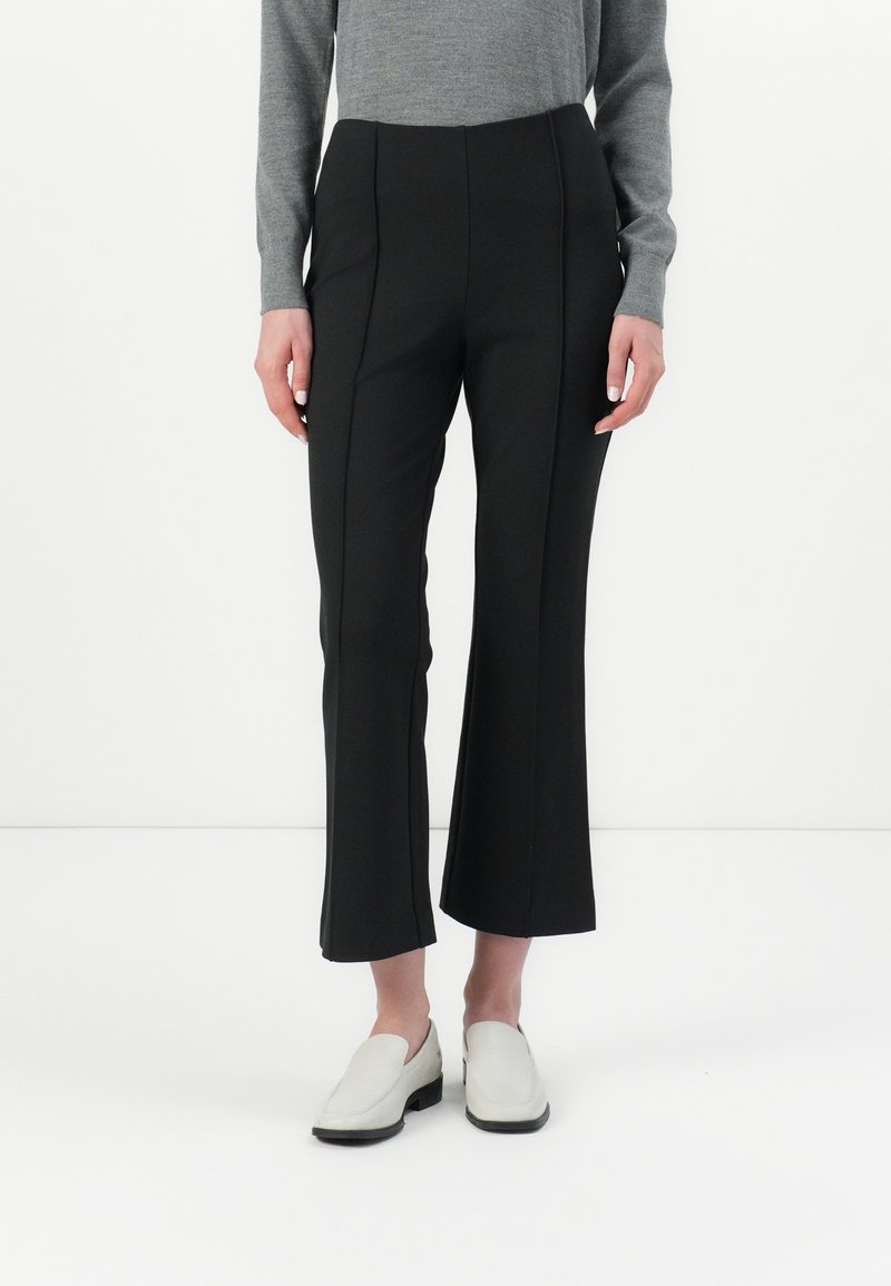 Personne portant un pantalon flare court noir, un haut gris à manches longues et des chaussures blanches slip-on, debout sur un fond blanc.