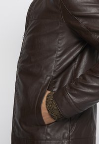 Chaqueta de cuero marrón con una textura suave, que presenta bolsillos laterales y un puño de punto en un color contrastante. Cierre de cremallera en la parte frontal.
