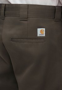 Καφέ παντελόνι από ανθεκτικό ύφασμα, με πίσω τσέπη και λευκή ετικέτα με πορτοκαλί λογότυπο. Η λεπτομέρεια περιλαμβάνει καθαρές ραφές και επίπεδη μέση.