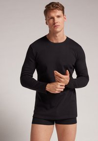 Zwarte langemouwen shirt met een ronde hals, gemaakt van een gladde stof. Model draagt zwarte onderkleding, wat een schone, minimalistische uitstraling laat zien.