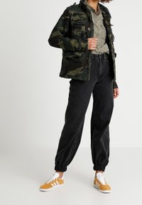 Kurtka kamuflażowa, zielona koszula zapinana na guziki, czarne, luźne spodnie oraz żółto-białe sneakersy. Posiada kieszenie, elastyczne mankiety oraz luźny krój.