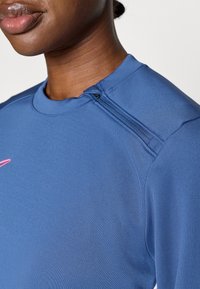 Μπλε αθλητική μπλούζα με λεία υφή, διαθέτει φερμουάρ στον ώμο και ροζ διακοσμητικές ραφές κατά μήκος του λαιμού.