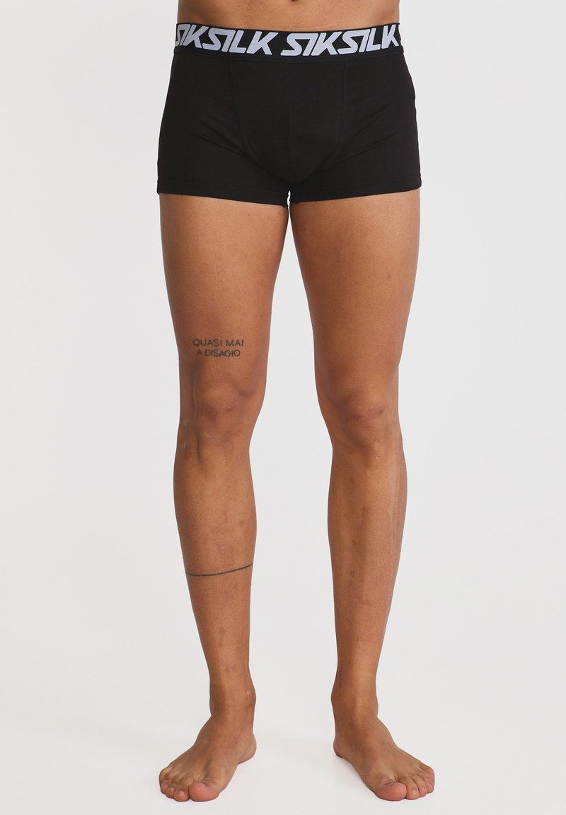 Άνδρας που φοράει μαύρα μποξέρ SikSilk, στέκεται ξυπόλητος μπροστά σε μονόχρωμο φόντο, με δύο μίνιμαλ τατουάζ στο αριστερό πόδι.