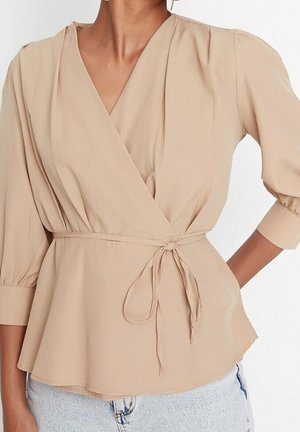 Vrouw draagt een beige wikkelblouse met 3/4 pofmouwen die in de taille is gestrikt, gecombineerd met lichtblauwe spijkerbroek.