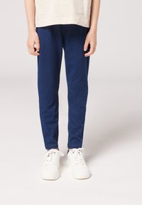 Námořnické modré slim fit kalhoty vyrobené z měkkého denimu, s hladkou texturou a minimálním zdobením. Vybavené bílými teniskami pro kontrast.