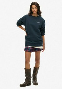 Sudadera azul marino con el logo de "Superdry", combinada con una minifalda de cuadros rojos y verdes y botas de rodilla marrones con tachuelas.