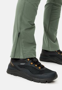 Groene technische broek met ritsen bij de enkels, gecombineerd met zwarte wandelschoenen met gaas- en rubberaccenten, oranje veters en logodetails.
