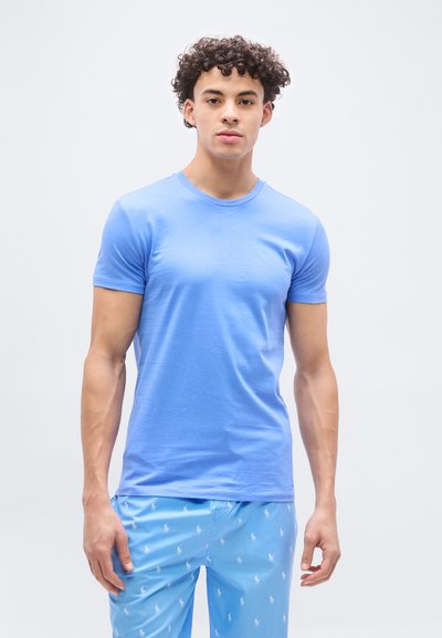 Νεαρός με κατσαρά μαλλιά, φορώντας ένα ανοιχτό μπλε κοντομάνικο πουκάμισο και μπλε σορτς με σχέδια, στέκεται μπροστά σε απλό λευκό φόντο.