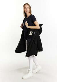 Giacca a vento nera con zip, tasca a toppa con un'illustrazione, indossata sopra una maglietta blu navy, abbinata a pantaloni bianchi e sneakers.