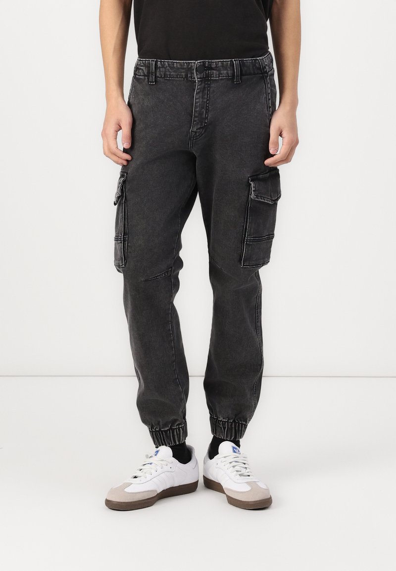 Cargo-pantalons in vervaagd zwart denim met elastische boorden, dubbele zijzakken en contrasterende witte stiknaad. Gedragen met witte sneakers.