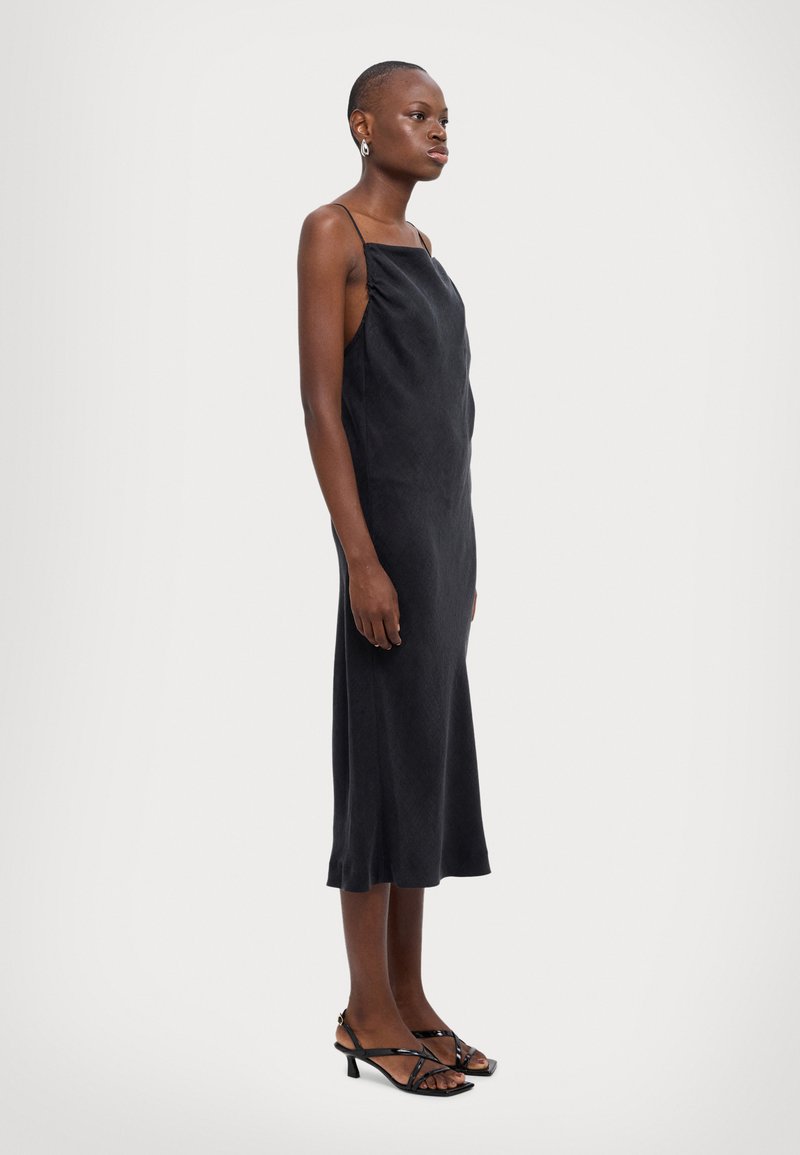 Black sleeveless dress with a relaxed fit, smooth fabric, mid-length, side slit, and minimal strap design. Worn with strappy black heels.